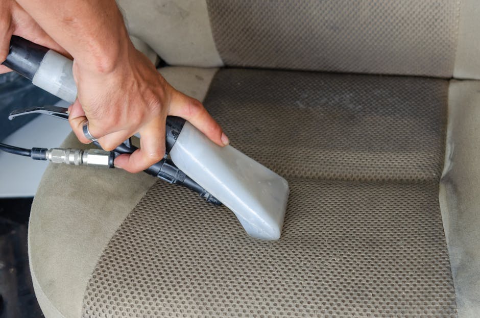 Close-up of a person using a handheld electric upholstery steamer on a dark grey fabric sofa in a living room, with visible cleaning motion on the seat cushion. The sofa has a modern design with a str